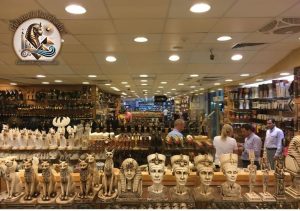 Interiorul unui magazin de suveniruri din Egipt, cu rafturi pline de statuete faraonice, pisici Bastet, papirusuri și turiști care aleg cadouri.