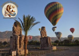 Vedere panoramică surprinsă într-o Excursie cu Balonul în Luxor, cu baloane multicolore zburând deasupra Coloșilor lui Memnon și a palmierilor.